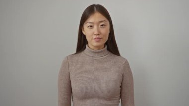 Boğazlı kazak giyen, beyaz bir arka plan karşısında gülümseyen bir kadın, basit ve neşeli bir ortamda pozitif bir jest ve ifade sergiliyor..