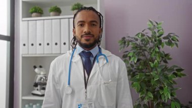 Doktor içeride steteskopla gülümsüyor. Klinikte dosyalar, bitki örtüsü ve profesyonel kıyafetlerle iyi bir jest yapıyor..