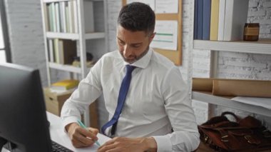 Ofiste çalışan bir adam masasında notlar yazıyor, etrafı kitaplar ve bilgisayarlarla çevrili, odaklanma ve üretkenliği vurgulayan profesyonel bir iş ortamı yaratıyor..