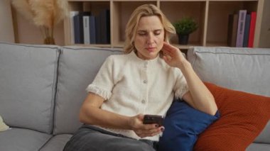 Kanepede oturan kadın rahat oturma odasındaki akıllı telefonu kullanıyor. Evin içi kitaplık arka planı ve bej dekoruyla dolu.