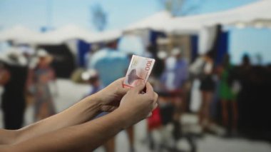 Norveç Kronu 'nu elinde tutan adam kalabalık bir pazar sokağında arka planda bulanık alışveriş merkezleri ve mağaza çadırları ile sokak ticaretini vurguluyor..