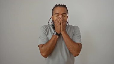 Örgülü genç adam esniyor ve beyaz bir duvara yaslanıyor, gri bir tişört giyiyor, sabah yorgunluğunu izole bir ortamda iletiyor..