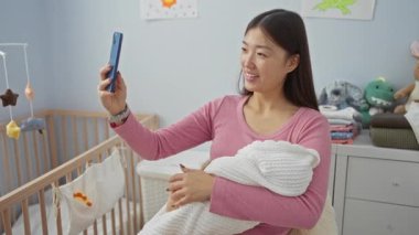 Bebek kucağındayken gülümseyen ve rahat bir yatak odasında beşiği, gösterişli anneliği ve modern teknolojisi olan selfie çeken bir kadın..