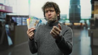 Kapalı alanda elinde İsviçre frangı olan bir adam havaalanı terminalinde kameraya doğrultuyor, elinde İsviçre parası olan ciddi bir ifade veriyor ve geçmişi bulanıklaştırıyor..