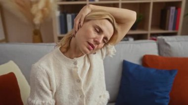 Mavi ve kırmızı yastıklı, rahat bir oturma odasında boynunu uzatan orta yaşlı ve sarışın bir kadın. Evdeki bir kanepede dinleniyor..