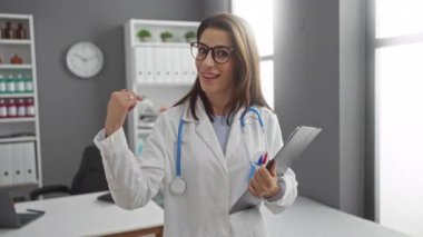 Klinikteki kadın gülümsüyor ve parlak ofis ortamında steteskop ve panoyla kendine güvenerek profesyonel ve olumlu ambiyansı vurguluyor..