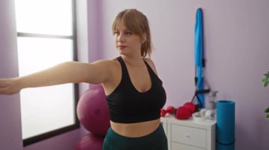 Spor malzemeleriyle spor salonunda esneyen, spor kıyafetleri giyen, spor toplarıyla çevrili, aydınlık, modern spor salonunda duran bir kadın..