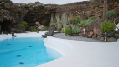 Lanzarote, İspanya 'da bir açık havuzun bulanık arka planı, dağılmış kayalık çevresi, bitkiler ve bokeh etkisi.