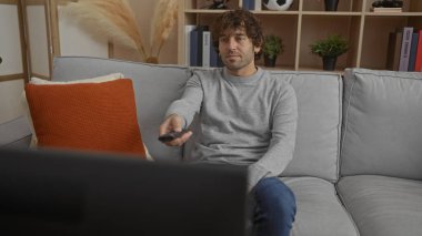 Oturma odasında televizyon izleyen genç bir adam, rahat, uzaktan kumandalı, rahat bir şekilde kanepede oturuyor, modern bir dairede şık bir dekoru var..