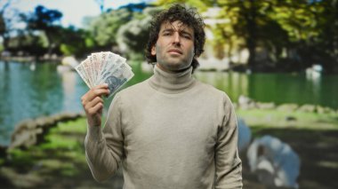 Elinde Polonya parası tutan İspanyol bir adam, arka planda sakin bir göleti olan canlı yeşil ağaçlarla çevrili yemyeşil bir parkta güvenle duruyor. Bu da açık havada finansal başarı olduğunu gösteriyor..