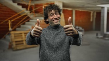 İnşaat alanında gülümseyen genç adam kentsel gelişimin ortasında güven ve pozitifliği temsil eden günlük kıyafetleri ve mutlu ifadeleri ile başparmağıyla onaylıyor..