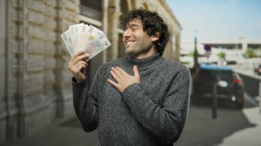 İspanyol adam açık bir gökyüzünün altında açık bir caddede dururken, Czech Koruna banknotlarını mutlulukla tutuyor, bir şehir ortamında neşe ve finansal başarı sergiliyor..
