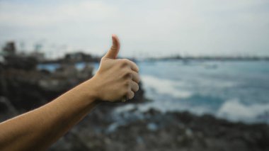 Arka planda bulanık okyanusla deniz kenarında baş parmak gösteren adam, suyun kenarındaki sakin sahil atmosferinde pozitifliği ve onayı tasvir ediyor..