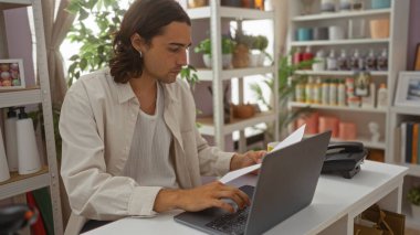 Etrafında çeşitli dekoratif eşyalar sergilenen bir mağazada dizüstü bilgisayarla çalışırken gazete okuyan uzun saçlı İspanyol bir genç.