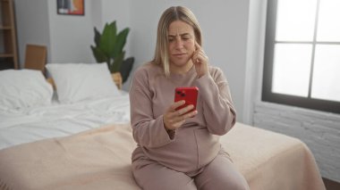 Sarışın hamile bir kadın, rahat bir yatak odasında, elinde akıllı bir telefon tutarak, evde huzurlu bir atmosfer yansıtıyor, evde bebek bekliyor..