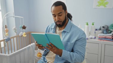 Yatak odasında beşik ve bebek eşyalarıyla kitap okuyan genç adam, aile ortamları için uygun rahat ve sakin bir ortam sergiliyor..