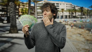 Güneşli bir sahil sahilinde Rus rublesi tutan İspanyol adam, para, seyahat ve beklenmedik zenginlik temaları çağrıştırıyor..