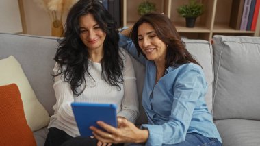 Olgun kadınlar oturma odasındaki kanepede birlikte vakit geçiriyorlar, tablet ekrana bakarken gülüyorlar, ev ve aile sevgisinin sıcaklığını kucaklıyorlar..