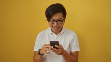 Genç Çinli adam, izole edilmiş sarı arka plana karşı akıllı telefon kullanıyor, gülümsüyor ve odaklanmış görünüyor.
