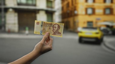 Bir şehir caddesinde, arka planda binaları ve arabaları olan, kentsel değişim manzarası sergileyen, Macar paralarını elinde tutan bir adam..