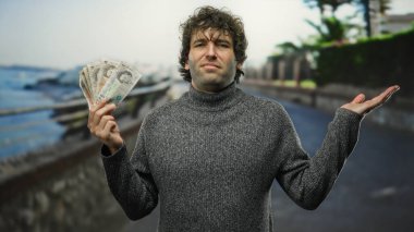 İspanyol asıllı bir adam, elinde İngiliz poundu ile sahil kıyısında duruyor, kendine güvenen bir ifadeyle, finans temalarını manzaralı kıyı titreşimleriyle harmanlıyor..