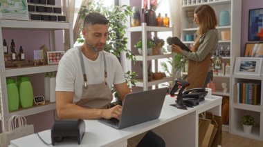 Kadın ve erkek, bir ev dekorasyonu mağazasında çalışıyor. Tezgahta dizüstü bilgisayar kullanan bir adam ve raflarda eşyalar düzenleyen bir kadın.