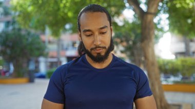 Sakallı genç bir adam arka planda ağaçlar ve binalarla şehir parkında dikiliyor. Mavi bir gömlek giyiyor. Düşünceli ve özenli görünüyor..