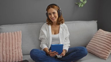 Oturma odasındaki kanepede oturan, kulaklık takan, tablet tutan, yastıklı ve bitkilerli rahat bir ortamda gülümseyen genç bir kadın..