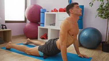 Spor salonundaki mavi bir paspasın üzerinde yoga yapan çekici, genç İspanyol bir adam. Toplar ve ağırlıklar da dahil olmak üzere spor aletleriyle çevrili. Sağlıklı bir yaşam tarzı gösteriyor..