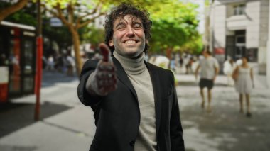 Canlı bir şehir sokak ortamında elini uzatarak gülümseyen genç İspanyol adam davetkar ve dost canlısı bir şehir atmosferini yakalıyor..