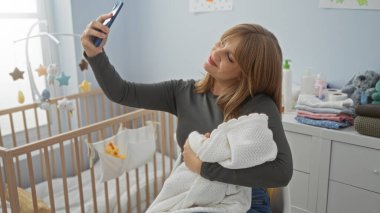 Bebek odasında, battaniyeye sarılı, bebek beşiği, pelüş oyuncaklar ve bebek malzemeleri ile çevrili bir şekilde selfie çeken kadın, neşeli bir iç mekan anı yakalıyor..