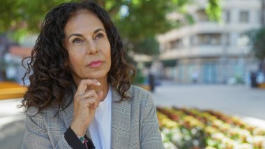 Çevresi yeşillik ve şehir manzarasıyla çevrili bir şehir parkında düşünceli bir ifade takınan, neşeli, güneşli bir günde bulanık şehir arka planına sahip bir kadın..