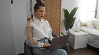 Konforlu yatak odasındaki dizüstü bilgisayarda çalışan bir kadın ev bitkileri ve modern dekorlarla günlük giysiler giyiyor, sakin bir ev ortamındaki görevlere odaklanıyor..