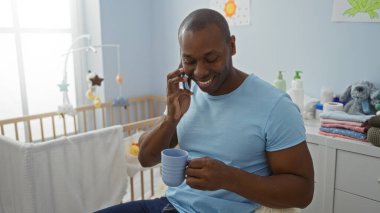 Yatak odasında gülümseyen genç bir adam telefonda konuşurken ve bardağı beşiğin yanında tutarken ev içinde sıcak ve davetkar bir atmosfer yaratıyor..