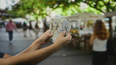 Kalabalık bir şehir caddesinde, gün ışığında ağaçlar ve insanlarla çevrili açık bir kafede Amerikan doları tutan bir adam..