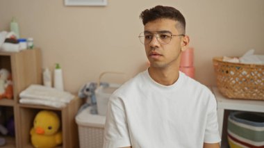 Gözlüklü genç adam modern ev içinde düşünceli görünüyor. Etrafı yumuşak oyuncaklar ve ev eşyalarıyla çevrili.