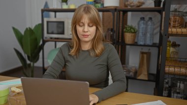 Ofisteki dizüstü bilgisayarında iç mekanlar, raflar ve modern dekorlarla çalışan kadın, üretkenlik ve estetiğe odaklı profesyonel bir ortam sergiliyor..