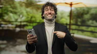 Açık havada gülümseyen ve kendine güvenen genç İspanyol bir adam bir park düzenine güvenerek, elinde akıllı bir telefonla modern yaşam tarzını sergiliyor, doğayla iç içe..