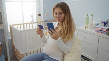 Hamile kadın yatak odasında oturmuş akıllı telefon kullanıyor ve online alışveriş sırasında bebek beşiğinin yanında kredi kartı tutuyor.