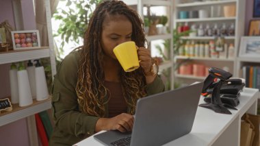 Ev dekorasyonu mağazasında dizüstü bilgisayarda çalışan bir kadın, çeşitli dekoratif eşyalar ve raflarla çevrili kahvesini yudumluyor..