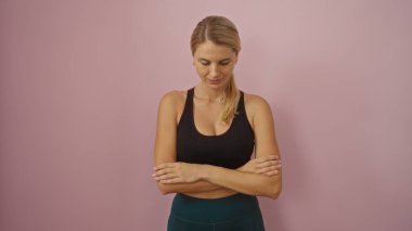 Pembe arka planda duran, kolları çaprazlanmış, aşağı bakan, siyah bluz giyen, rahat duruş takınan, sarı saçlı, kendine güvenen bir kadın, izole edilmiş bir ortam..