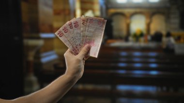 Kapalı bir kilise ortamında Türk lirası tutan adam, dini bir ortamda paranın zıtlığını vurguluyor, renkli banknotlarla yakın plan çekimleri yapıyor..