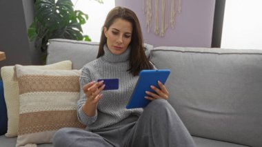 Oturma odasında elinde kredi kartı ve tabletle alışveriş yapan bir kadın modern teknoloji kullanımını öneren rahat bir ev ortamını öneriyor..