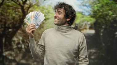 Güney Kore 'nin paralarını bir park ortamında tutan gülümseyen bir adam, doğanın ortasında finansal başarı ve kültürel çeşitlilik sergiliyor..