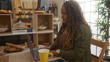 Samimi bir fırında dizüstü bilgisayar başında çalışan bir kadın. Etrafı ekmek ve hamur işleriyle çevrili. Kapalı bir kafedeki masada sarı bir kahve fincanı var..
