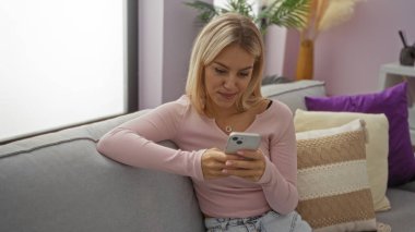 Kadın rahat bir oturma odasında, rahat bir kanepede rahat bir şekilde oturuyor, akıllı bir telefon kullanıyor, ev dekoru unsurları ile modern bir hayat ve rahatlık hissi uyandırıyor..