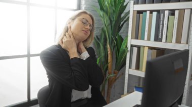 Kadın, arka planda kitaplıklar ve bitkilerle dolu modern bir ofiste masa başında otururken boyun ağrısı çekiyor. Tipik bir çalışma ortamı olduğunu gösteriyor..