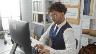 Modern ofis ortamında bilgisayarda çalışan, gözlük ve yelek takan genç İspanyol bir adam, iyi organize edilmiş bir çalışma alanı içindeki görevine odaklanmış..