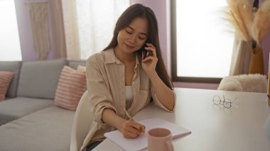 Evinde rahat bir oturma odasında notlar alan Çinli bir kadın telefonda konuşuyor..