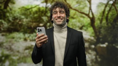 Parkta, arka planda ağaçlarla çevrili bir parkta akıllı telefon kullanarak gülümseyen genç adam yakışıklı ve kendinden emin bir ifade sergiliyor..